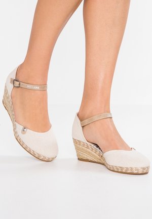 Pieds chaussés d'espadrilles compensées beige avec des brides à la cheville et des semelles en jute tissé sur fond blanc.