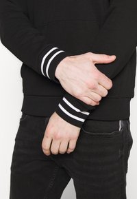Černá mikina s bílými pruhovanými náplety, se spodní kapsou a uvolněným střihem. Nosit s tmavými džíny, ruce sepnuté kolem paží.