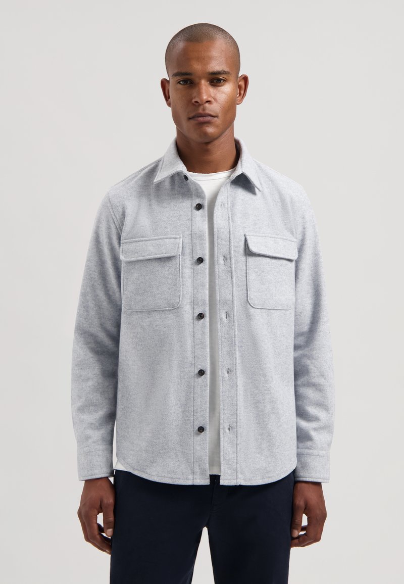 Szara wełniana koszula z dwoma kieszeniami na piersi, zapięciem na guziki z przodu i kołnierzykiem. Koszula ma miękką teksturę i zaokrąglony dół.
