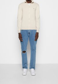 Persona che indossa un maglione crema con cerniera, jeans azzurri strappati con un buco al ginocchio e scarpe da ginnastica bianche, in piedi contro uno sfondo bianco.