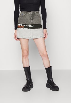Kobieta nosząca mini spódniczkę dżinsową w blokach kolorystycznych z napisem "KARL LAGERFELD JEANS", czarną bluzkę z długim rękawem i czarne skórzane botki za kostkę.