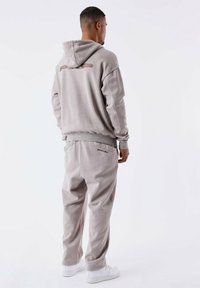 Lichtgrijze hoodie en bijpassende joggingbroek van zachte stof. De hoodie heeft een logo op de rug; de broek heeft zijzakken. Witte sneakers maken de outfit compleet.