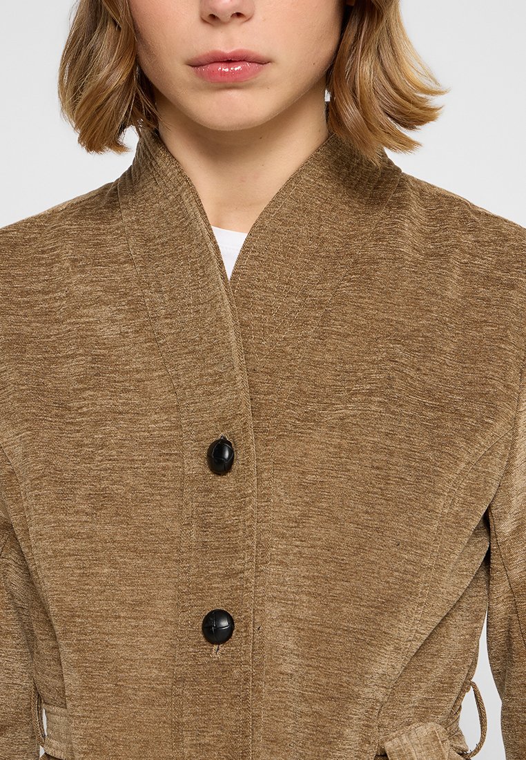 Giacca marrone con texture, caratterizzata da un alto colletto, tre bottoni neri e un nastro in vita. Il tessuto ha un aspetto morbido e informale.