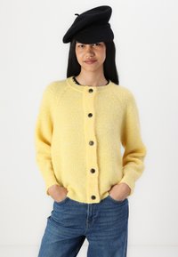 Gilet en laine jaune avec un col arrondi, fermeture à boutons et texture côtelée, associé à un jean en denim bleu et un béret noir.