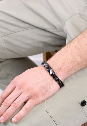 Schwarzes Lederarmband mit einem geflochtenen Design und einem glatten metallischen Akzent, der geometrische Formen aufweist, am Handgelenk getragen.
