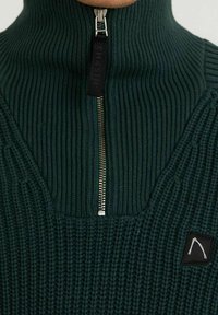 Tmavozelený pletený svetr s vysokým límcem, se zapínáním na přední straně a malou černou nášivkou na levé straně. Celoplošný texturovaný vzor.