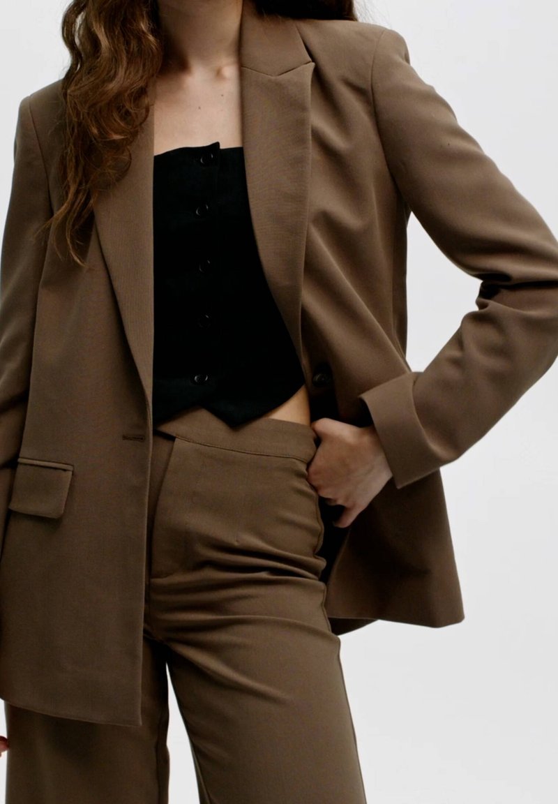 Blazer marron sur mesure avec épaules structurées, bouton unique, associé à un haut noir ajusté et un pantalon taille haute assorti. Texture lisse.