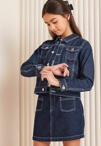 Temno modra jeans jakna z belim šivom, sprednjimi žepi in gumbnim zapiranjem, v kombinaciji z ujemajočo se jeans krilom, ki ima žepa.