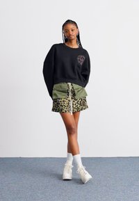 Čierna oversized mikina s grafikou srdca, vrstvená s našuchovanou zelenou sukňou s leopardím vzorom. Biely tenisky a športové ponožky dotvárajú celkový vzhľad.