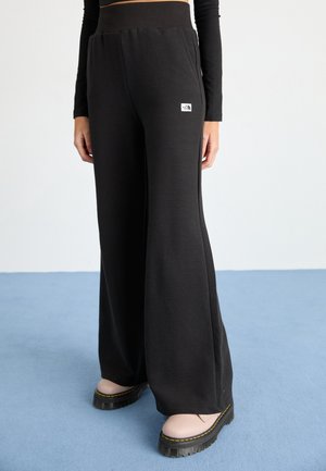 Person trägt schwarze, weit geschnittene Hose mit The North Face-Logo, schwarzes Oberteil und hellrosa Plateauschuhe auf blauem Boden.