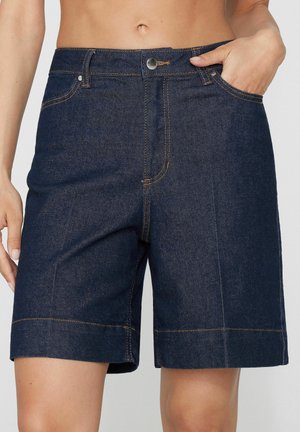 Dunkelblaue Jeansshorts mit orangefarbenen Nähten, vorderem Knopfverschluss und Taschen; getragen von einer Person mit einer Hand in der Tasche.