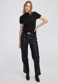 Kobieta stojąca z jedną ręką na biodrze, ubrana w czarną bluzkę z krótkim rękawem, czarne skórzane spodnie z paskiem i czarne botki na obcasie, na jednolitym tle.