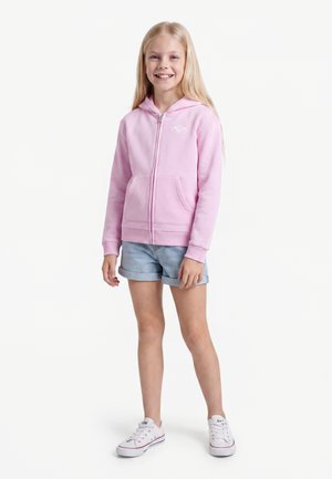 Fille souriante aux longs cheveux blonds portant un sweat à capuche rose zippé, un short en jean bleu clair retroussé et des baskets blanches, debout face à nous.