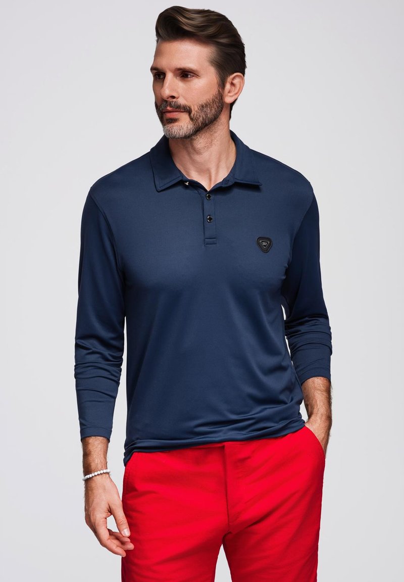 Polo a maniche lunghe blu navy in tessuto liscio con colletto a bottoni e un piccolo logo nero sul petto, abbinato a pantaloni rossi.