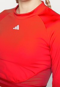 Czerwona sportowa koszulka z długimi rękawami, okrągłym dekoltem, gładką fakturą oraz białym logo na piersi. Posiada dopasowany krój.