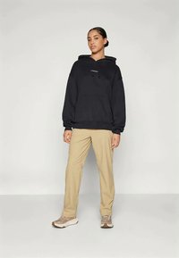 Mujer de pie, vistiendo una sudadera negra, pantalones beige y zapatillas beige, contra un fondo claro y liso.