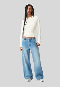 Langærmet hvid top med skulderudskæringer og dekorative øjer, parret med vide lyseblå jeans. Glat stof og afslappet pasform.