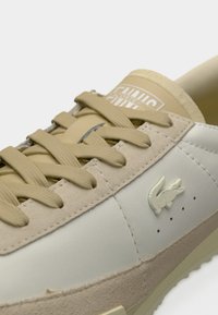 Μπεζ και ανοιχτό γκρι αθλητικό παπούτσι με υφασμάτινα κορδόνια, σουέντ λεπτομέρειες και λογότυπο κροκόδειλου στην πλευρά κοντά στο μπροστινό μέρος.