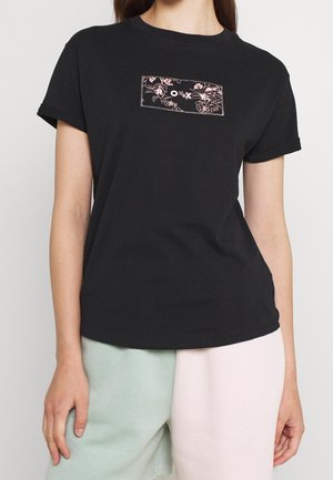 Žena v černém triku s krátkými rukávy, na kterém je růžový květinový obdélníkový motiv a uprostřed na hrudi jsou písmena "O X".