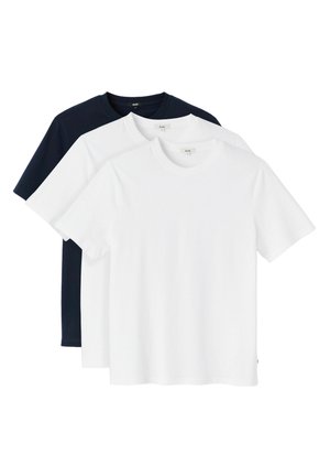 Jeune homme portant un t-shirt blanc, un jean bleu clair et une casquette de baseball lavande portée à l'envers, debout devant un fond uni.