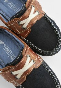 Marineblaue Wildleder-Bootsschuhe mit braunen Lederakzenten, weißen Schnürsenkeln und kontrastierenden Nähten. Strukturierte Oberfläche und gepolsterte blaue Innensohle.