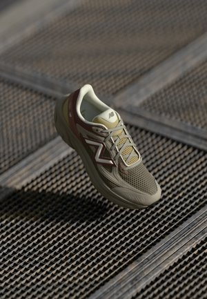 Lichtbruine en beige New Balance sneaker met mesh- en suède details, rustend op een gestructureerde metalen rooster vloer.