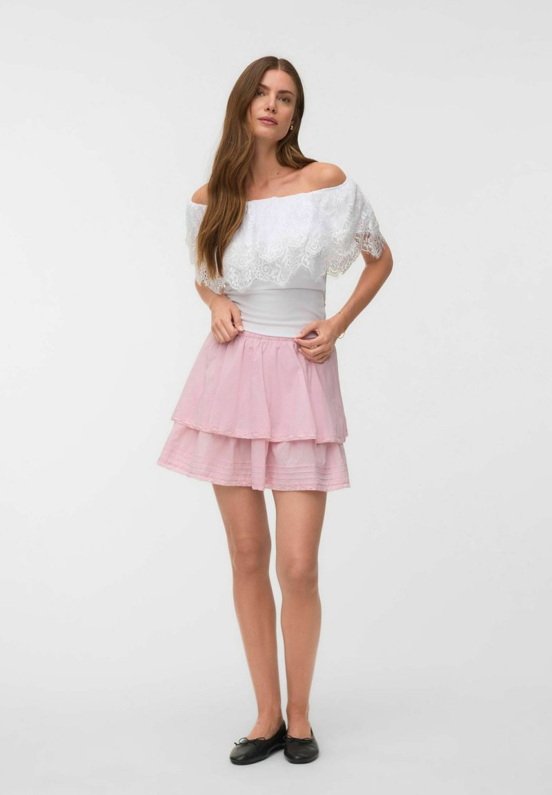 Femme aux longs cheveux bruns portant un top en dentelle blanc à épaules dénudées, une jupe rose à volants et des ballerines noires, debout sur un fond blanc.