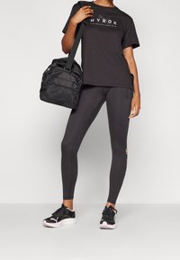 Camiseta deportiva negra con logo blanco, combinada con leggings oscuros que tienen un acento amarillo. Zapatillas negras y un bolso de deporte negro con detalles de malla.