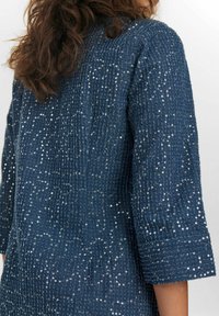 Top blu navy con paillettes, con finitura testurizzata e maniche a tre quarti. Il tessuto presenta un motivo di paillettes scintillanti su tutta la superficie.