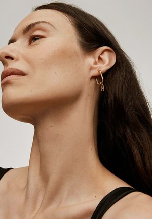 Pendiente de aro en tono dorado con un pequeño charm colgante en forma de la letra "H". Diseño simple y textura de superficie lisa.