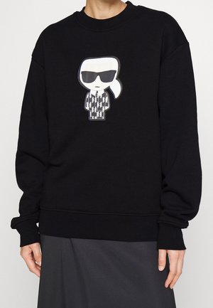 Czarna bluza z kapturem z grafiką kreskówkową przedstawiającą osobę w okularach przeciwsłonecznych i wzorzystym stroju, umieszczoną na środku klatki piersiowej.