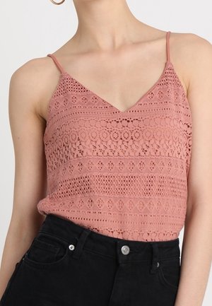 Kobieta w koronkowej bluzce na cienkich ramiączkach w kolorze pudrowego różu, wsuniętej w czarne, wysokowalne jeansy.