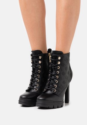 Mustat korkokengät, joissa on nyöritys ja kohokuvioidut kuviot, jalassa paljain jaloin, valkoista taustaa vasten.