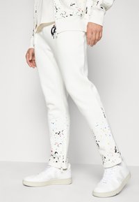 Hvite sweatpants med farge sprut mønstre og sidelommer, kombinert med hvite sneakers. Har en avslappet passform og mykt materiale.