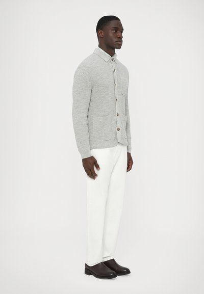 Pilka megzta kardiganinė striukė su apykakle, mediniais mygtukais ir dviem priekinėmis kišenėmis, derinama su baltais kelnėmis ir rudais batais.