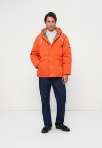 Orange padded jacket featuring a hood, white drawstrings, snap buttons, and side pockets. Worn over a striped shirt and dark trousers.