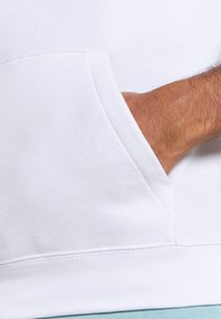 Main partiellement insérée dans la poche avant d'un sweat-shirt blanc, montrant les coutures de la poche et la texture du tissu.