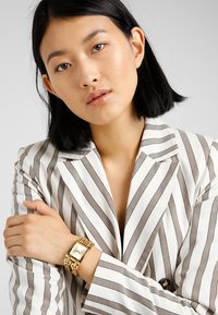 Gold-tone watch with square face and chain-link strap, worn on a striped blazer with beige and white vertical lines. Smooth texture.