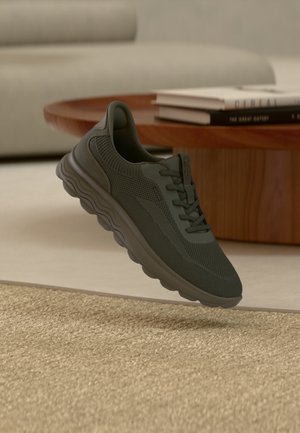 Zapatilla de running de malla gris oscuro con suela gruesa flotando sobre una alfombra tejida beige en una sala de estar con una mesa de café de madera y libros.