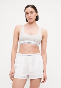 Svijetlosiva sportska grudnjak s teksturiranim uzorkom i širokom elastičnom trakom na kojoj je logo "Calvin Klein". U paru s bijelim, širokim kratkim hlačama.