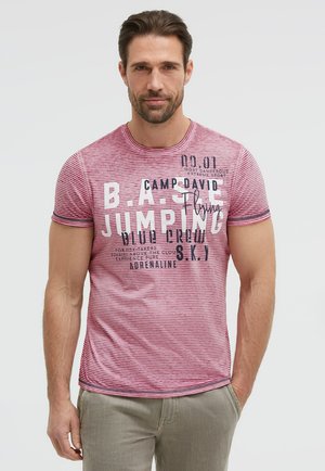 Mann mit kurzen braunen Haaren und Bart trägt ein rot gestreiftes T-Shirt mit Text und beigen Hosen, steht vor einem schlichten Hintergrund.