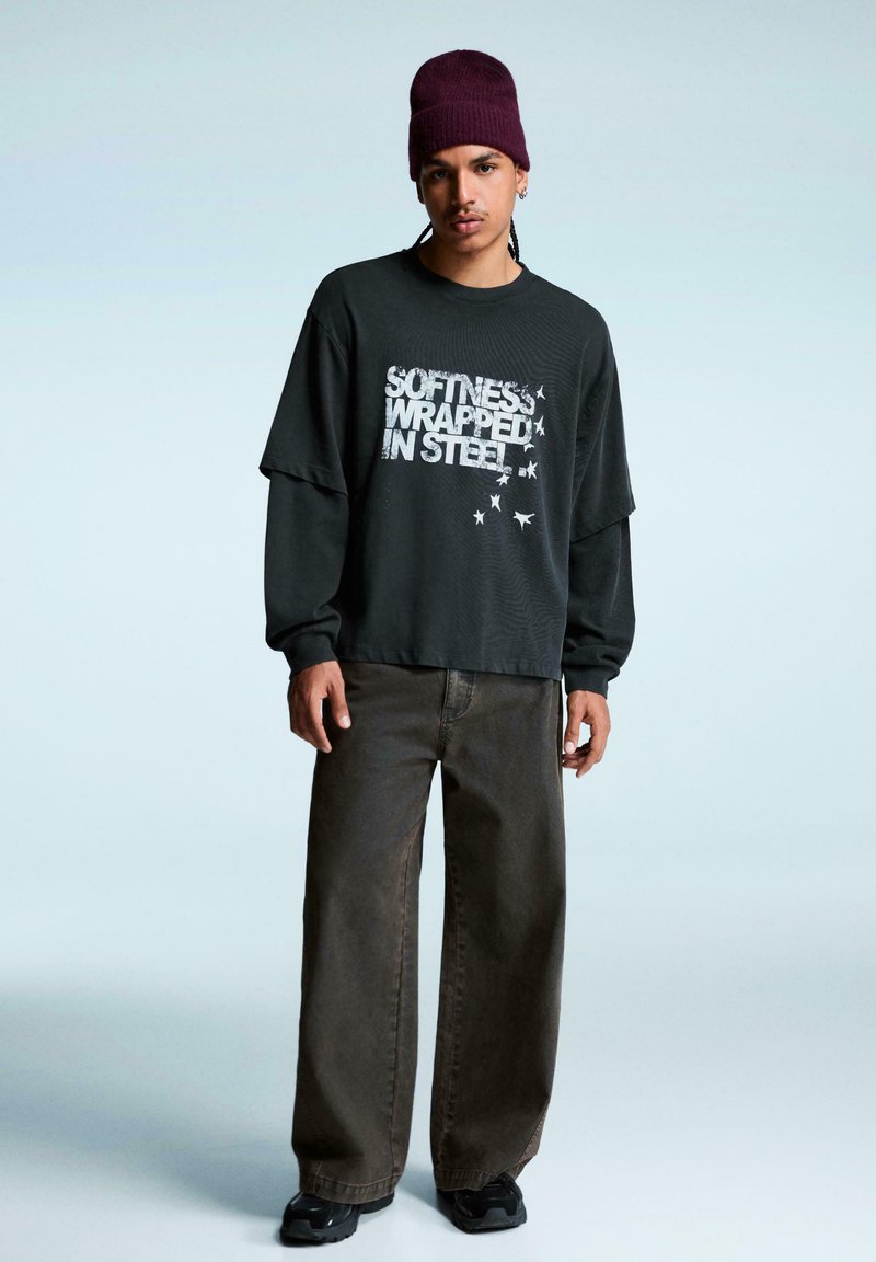 Fekete grafikás, hosszú ujjú póló "Puha acélban" felirattal, laza sötétbarna nadrággal és fekete sportcipővel párosítva; a modellen bordó sapka van.