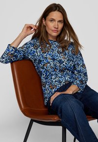 Blusenshirt mit blauem Muster und 3/4-Ärmeln, das florale Designs und kontrastierende orangefarbene Akzente aufweist, kombiniert mit dunklen Jeans aus Denim und vor einem braunen Stuhl positioniert.