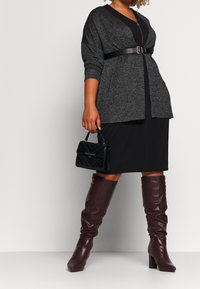 Cardigan grigio oversize con cintura, vestito nero aderente, stivali marroni fino al ginocchio e una piccola borsa nera con texture.