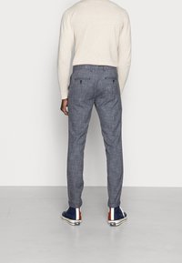 Šedé kostkované kalhoty s přizpůsobeným střihem, které mají dvě zadní kapsy a hladkou texturu. Nosené s modrými teniskami a světlejším svetrem.