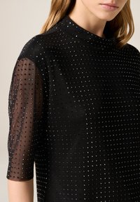 Czarny top z półdługimi rękawami, wykonany z gładkiej tkaniny z wypukłymi kropkami i wbudowanymi błyszczącymi elementami. Gładka faktura z wysokim dekoltem.