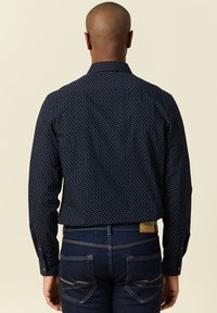 Camisa de botones azul marino con un pequeño patrón de puntos blancos, de mangas largas y con cuello puntiagudo. Combinada con jeans azul oscuro, que muestran un detallado cosido.