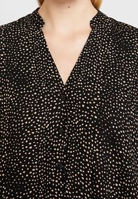 Czarna koszula z dekoltem w serek, z rozproszonym wzorem beżowych kropek oraz pięcioma czarnymi guzikami z przodu. Miękka faktura materiału.