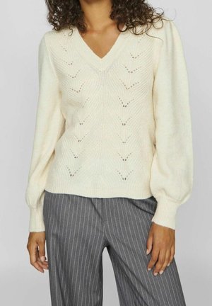 Vrouw die een crème-kleurige gebreide V-hals trui met een chevronpatroon en grijze gestreepte broek draagt, staand tegen een lichte achtergrond.