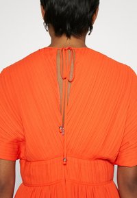 Osoba ubrana w jasnopomarańczową plisowaną sukienkę z krótkimi rękawami, z wycięciem z tyłu zapinanym na dwie cienkie sznurki zakończone małymi metalowymi koralikami.